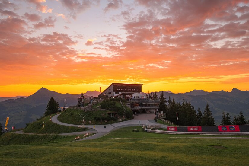 Berghütte auf einem Gipfel vor einem orangefarbenen Sonnenaufgang in den Alpen.