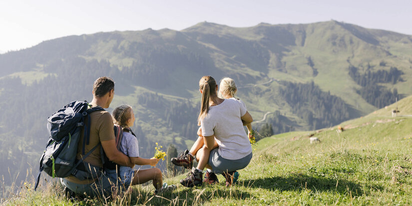 Familie sitzt auf einer Wiese und genießt die Bergsicht mit Blumen in der Hand in Kitzbühel.