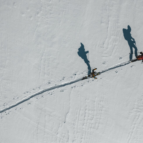 Zwei Personen auf Skiern durchqueren eine schneebedeckte Fläche, ihre Schatten zeichnen sich klar auf dem weißen Hintergrund ab.