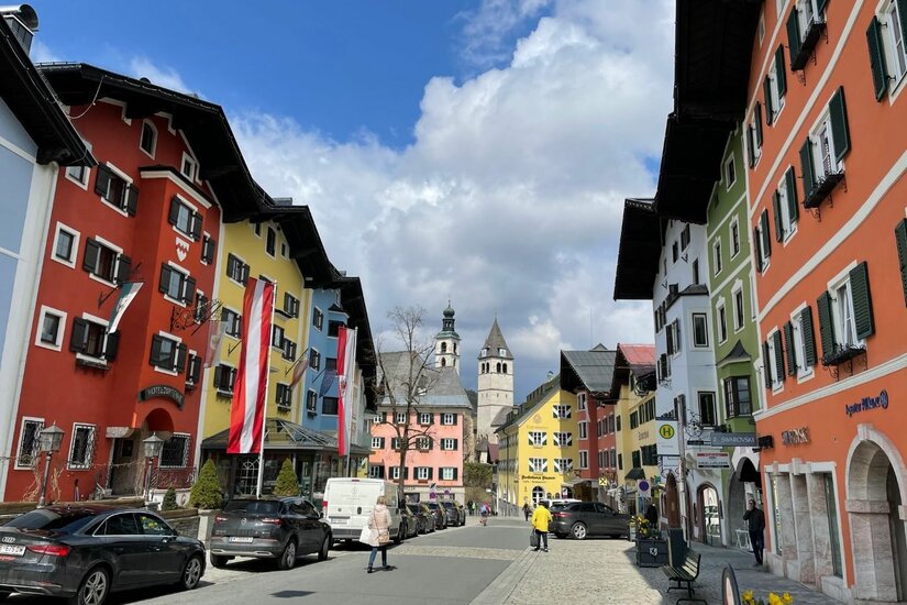 Blick durch eine Straße mit bunten Häuserfassaden und Kirchtürmen im Hintergrund unter bewölktem Himmel.
