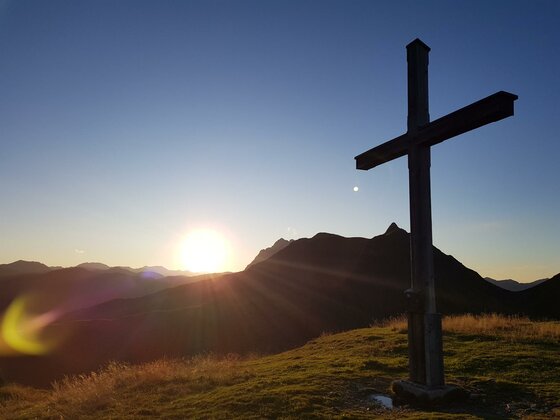 Gipfelkreuz bei der Sonnalm Ein hölzernes Gipfelkreuz auf einer Wiese vor einer Bergkulisse bei Sonnenuntergang.