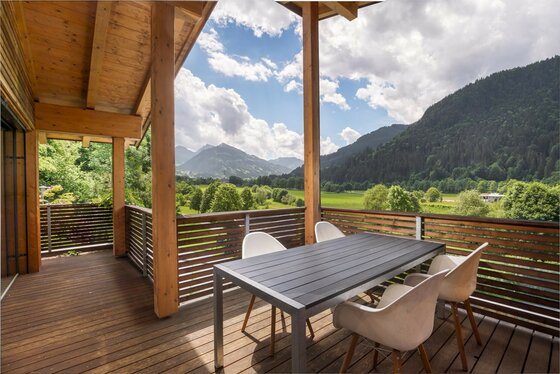 Balkon mit Bergblick Holzbalkon mit Sitzgruppe und Blick auf bewaldete Berge und grüne Wiesen unter bewölktem Himmel.