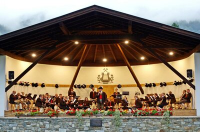 Eine Musikkapelle spielt auf einer Bühne in einer ansprechenden Umgebung. Im Hintergrund sind die Berge und ein bewölkter Himmel zu sehen.
