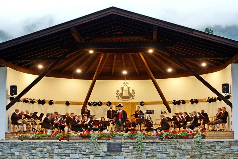 Pavillon Jochberg Eine Musikkapelle spielt auf einer Bühne in einer ansprechenden Umgebung. Im Hintergrund sind die Berge und ein bewölkter Himmel zu sehen.