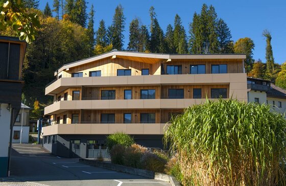 Mehrstöckiges Apartmenthaus mit Holzfassade und Balkonen vor einem bewaldeten Hang.