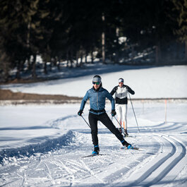 During a cross-country skiing event in Kitzbühel, two skiers glide along a snow-covered trail, surrounded by dark trees under a clear sky.