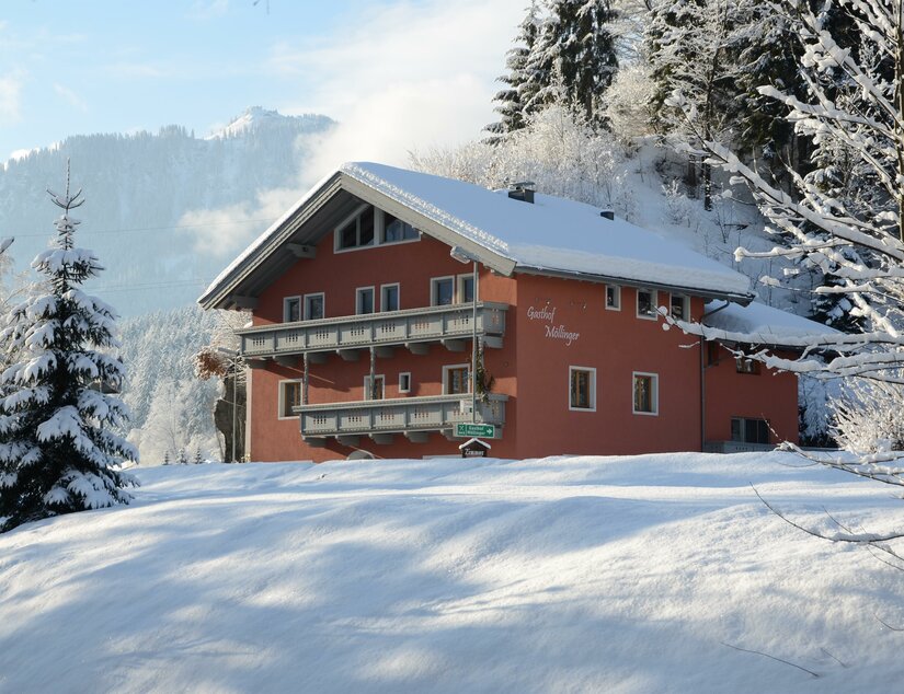 Ein rotes, schneebedecktes Gasthaus mit Holzbalkonen in einer winterlichen Berglandschaft.