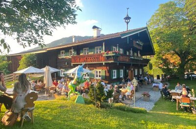 Traditionelles Holzhaus mit Außengastronomie auf einer grünen Wiese unter blauem Himmel.