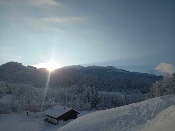 20231203_085640 Eine winterliche Landschaft mit schneebedeckten Bergen und einem Sonnenaufgang. Im Vordergrund steht ein kleines, eingeschneites Haus.