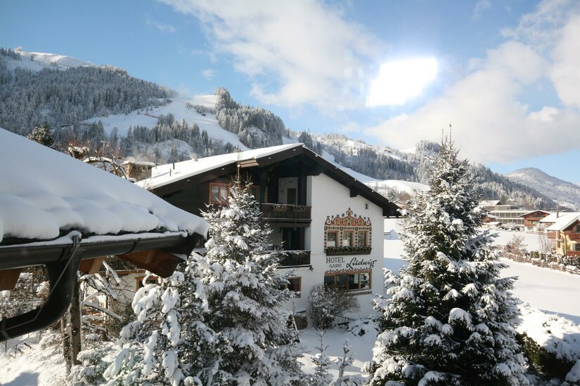 Verschneites Hotel Garni Ludwig vor bewaldeten Berghängen im Winter.