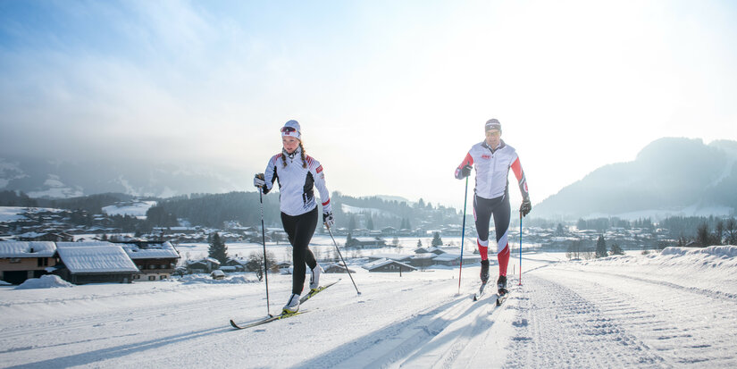 Two cross-country skiers glide along a snow-covered track, surrounded by a picturesque winter landscape in Kitzbühel.