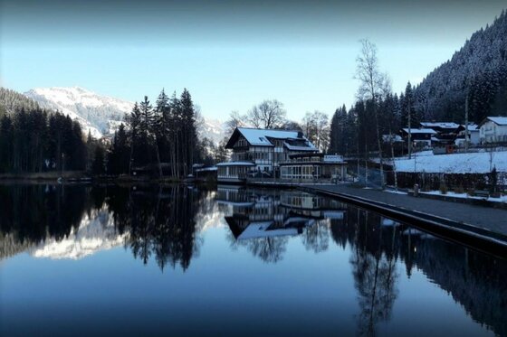 Pipino's im Winter Ein Gebäude am Seeufer mit Spiegelung im Wasser vor verschneiten Bergen und Nadelwald.
