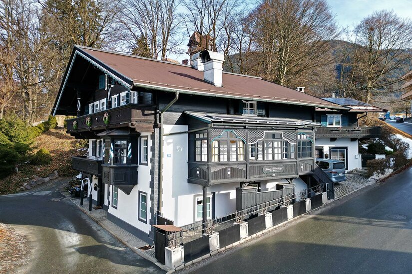 Hausansicht Hornbichl Traditionelles Alpenhaus mit dunkler Holzfassade, weißem Erdgeschoss und verzierten Balkonen vor bewaldetem Hang.