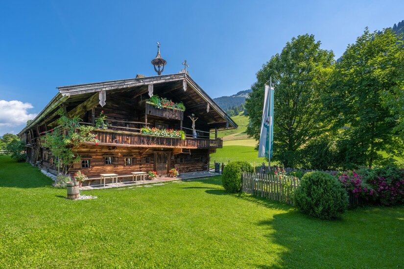Traditionelles Bauernhaus aus dunklem Holz mit Blumenschmuck auf einer grünen Wiese unter blauem Himmel.