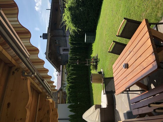Terrasse mit Holztisch, Stühlen und Markise vor einem Garten mit Rasenfläche und Hecke.