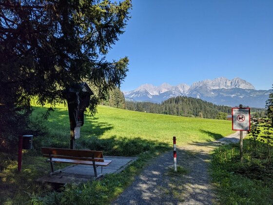 Wanderweg mit Holzbank, Marterl und Blick auf ein Gebirgsmassiv unter blauem Himmel.