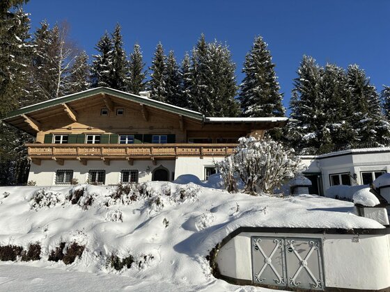 Alpines Holzhaus mit Balkon und weißer Fassade in einer verschneiten Winterlandschaft vor Nadelbäumen.