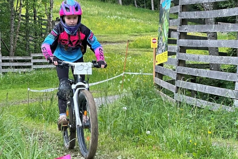 Ein Kind mit Helm und Schutzausrüstung fährt auf einem Mountainbike einen schmalen Schotterweg bergab.