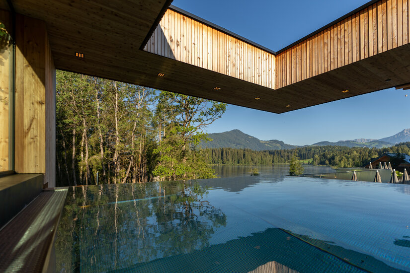 Ein moderner Pool mit glattem Wasser spiegelt die umliegende Natur und die Holzarchitektur unter einem klaren blauen Himmel wider.