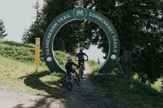 Start des Hahnenkamm Trails Zwei Mountainbiker fahren durch ein kreisförmiges grünes Portal auf einen Waldweg.
