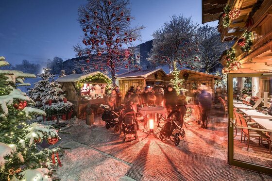 Beleuchteter Weihnachtsmarkt am Rasmushof bei Dämmerung mit Holzhütten, geschmückten Bäumen und Besuchern im Schnee.