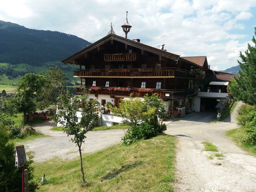 Großes, mehrstöckiges Holzhaus im alpinen Stil mit Blumenbalkonen vor bewaldeten Bergen.