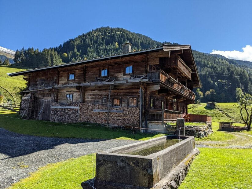 Jochberger 7-Brunnenweg Ein traditionelles Holzhaus in den Alpen mit einem steinernen Brunnen im Vordergrund.