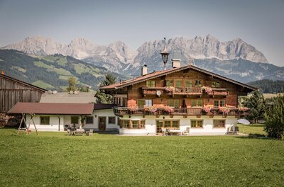 Alpines Holzhaus mit Blumenbalkonen vor einem schroffen Gebirgsmassiv.