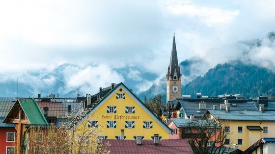 Stadtansicht Kitzbühel im Frühling Blick auf Kitzbüheler Häuserzeile mit dem gelben Hotel Tiefenbrunner und Kirchturm vor nebligen Bergen.