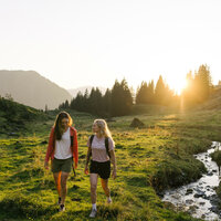 Zwei Frauen wandern fröhlich auf einer grünen Wiese, umgeben von Bergen und Bäumen, während die Sonne untergeht.