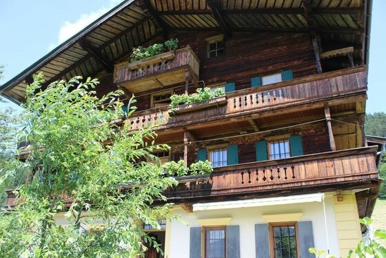 Ansicht Sommer Grünberghof Mehrstöckiges Holzhaus mit Balkonen, Blumenkästen und grünen Fensterläden vor blauem Himmel.