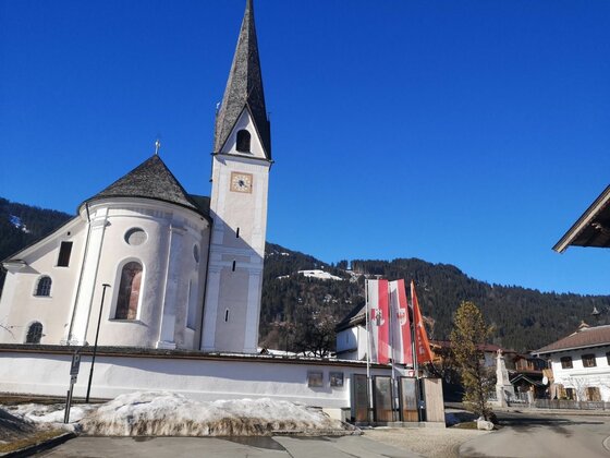 Kirche am Achensee Weiße Kirche mit spitzem Turm vor bewaldeten Bergen unter blauem Himmel.