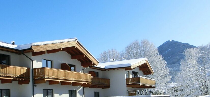 Alpines Gebäude mit Holzbalkonen und schneebedeckten Dächern vor blauem Himmel und Bergen.