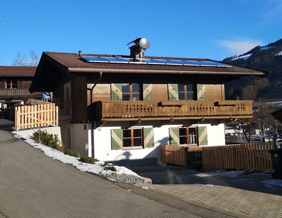 Zweistöckiges Alpenhaus mit Holzbalkon, grünen Fensterläden und Solaranlage auf dem Dach unter blauem Himmel.