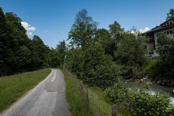 Asphaltierter Wanderweg neben einem Bachlauf und Bäumen unter blauem Himmel.