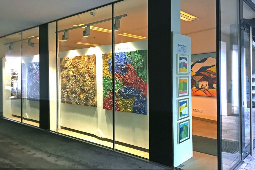 Eine Kunstgalerie mit großen, farbenfrohen Gemälden an der Wand. Die Fenster sind klar, und es gibt zusätzliche Kunstwerke in Rahmungen auf der rechten Seite.
