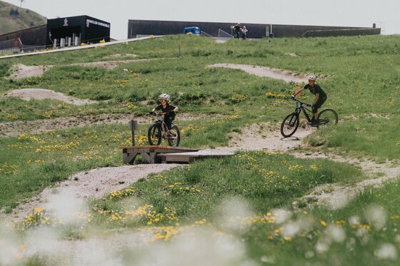 Zwei Mountainbiker auf einer Übungsstrecke mit Holzelementen und Kurven auf einer grünen Wiese.