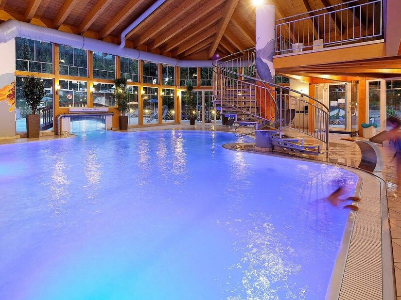 Ein modernes Hallenbad mit leuchtend blauem Wasser und einer Rutsche. Große Fenster sorgen für viel Tageslicht und eine entspannende Atmosphäre.