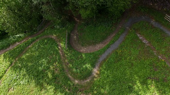 Mocking Uphill Trail Kurven Luftaufnahme eines geschwungenen Mountainbike-Uphill-Trails auf einer grünen Wiese am Waldrand.