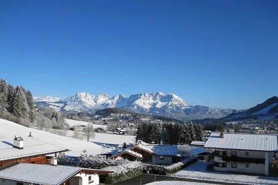 Verschneite Häuser im Vordergrund vor einem massiven, schneebedeckten Gebirgszug unter blauem Himmel.