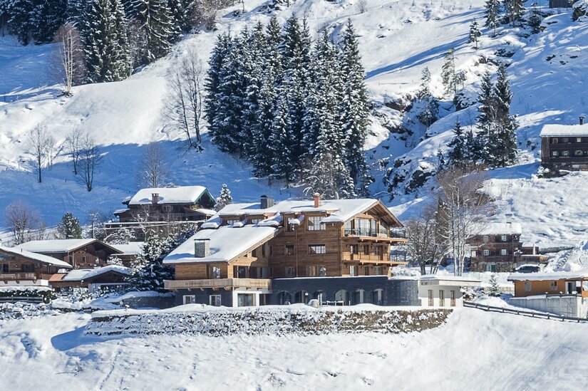 Mehrstöckiges Holzchalet mit schneebedecktem Dach in einer alpinen Winterlandschaft vor bewaldetem Hang.