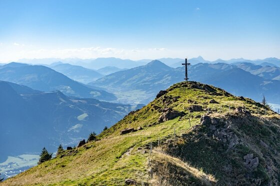 Gipfelkreuz am Stuckkogel Ein hölzernes Gipfelkreuz auf einem grasbewachsenen Bergrücken vor einem Panorama aus blauen Gebirgsketten.