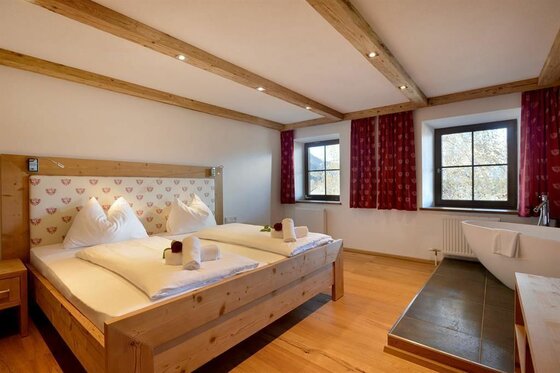 Schlafzimmer mit Doppelbett aus Holz, Holzbalkendecke und einer freistehenden Badewanne am Fenster.