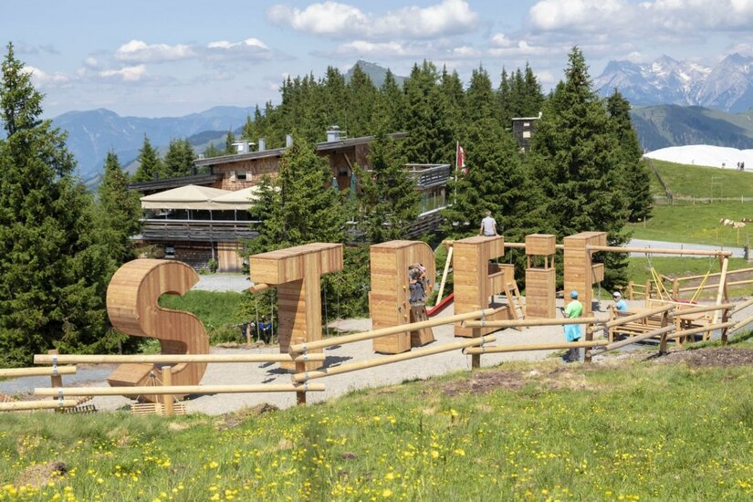 Sommererlebnis Streif Große Holzbuchstaben des Wortes STREIF als Spielstationen auf einer Almwiese vor einer Berghütte.