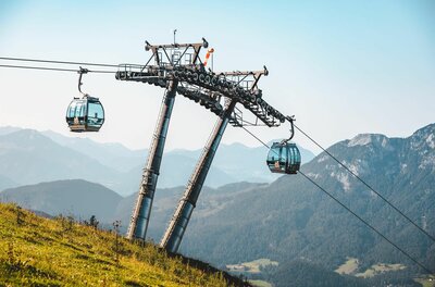 Eine Seilbahn mit blauen Gondeln schwebt über eine grüne Wiese. Im Hintergrund sind majestätische Berge und ein klarer Himmel zu sehen.