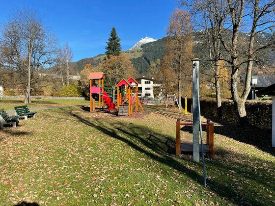 Kinderspielplatz mit Klettergerüst, Rutsche und Schaukeln vor bewaldeten Bergen unter blauem Himmel.