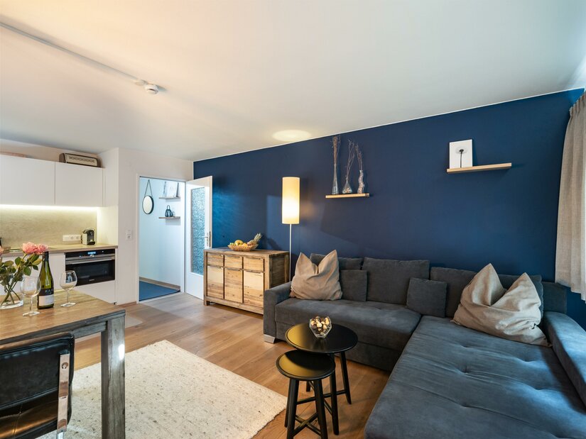 Modernes Wohnzimmer mit blauem Ecksofa, Holztisch und Küchenzeile im Hintergrund.