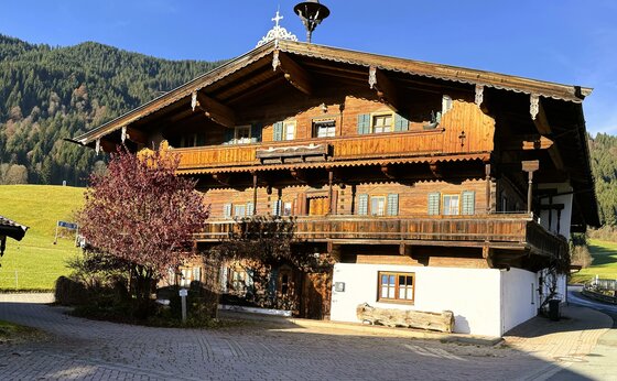 Ein traditionelles Holzhaus in malerischer Umgebung. Im Hintergrund erheben sich grüne Hügel und Bäume.