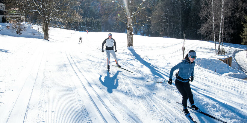 In this cross-country skiing scene in Kitzbühel, two skiers glide through a snow-covered landscape bathed in bright sunlight, with shadows cast across the landscape.