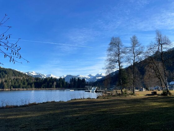 Schwarzsee Städtisches Freibad Blick über einen See auf schneebedeckte Berge unter blauem Himmel mit Bäumen im Vordergrund.